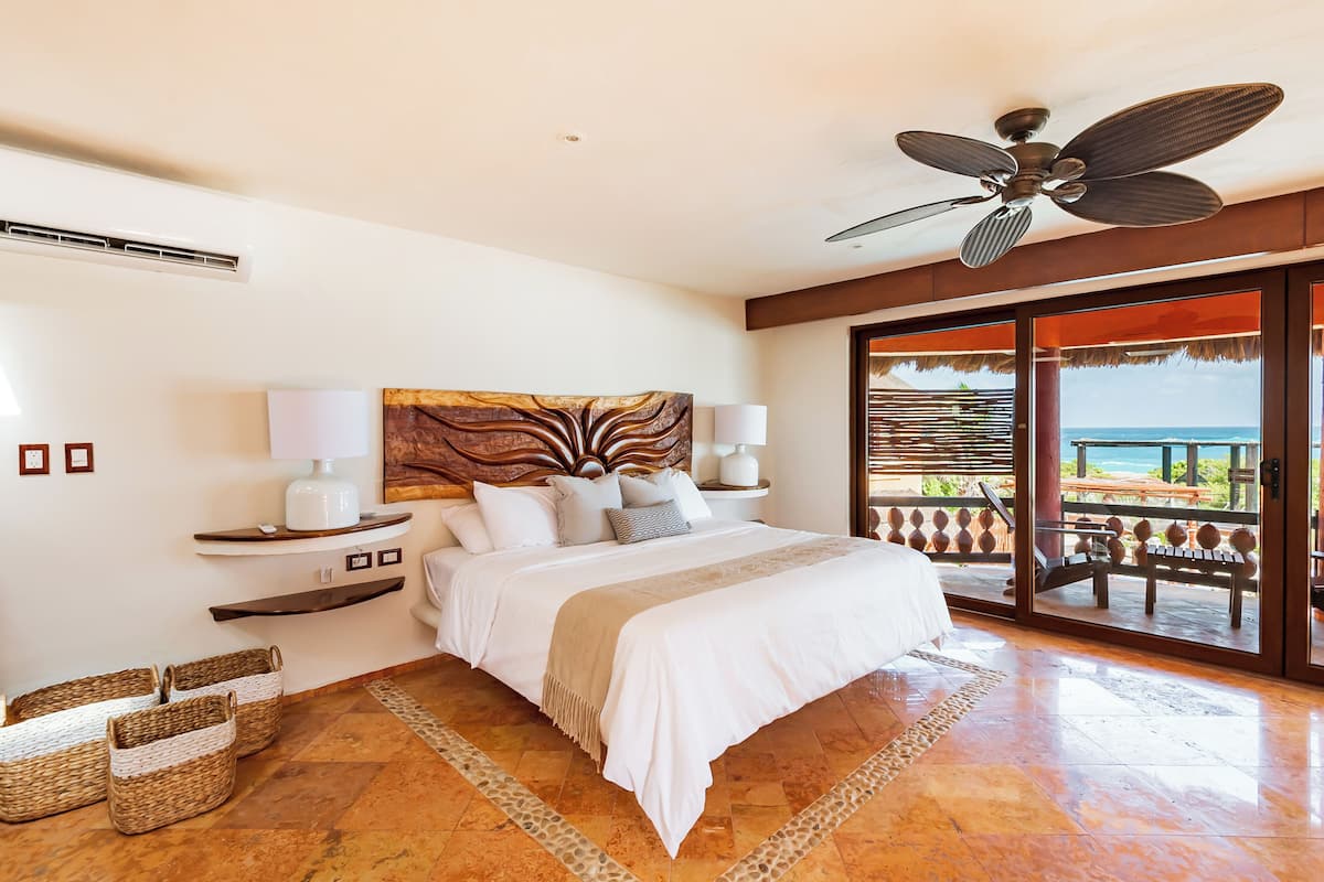A spacious bedroom with a large bed, a ceiling fan, a view of the ocean, and a balcony with outdoor seating.