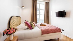 Ein Schlafzimmer mit einem Bett, einem Nachttisch mit Blumenvase, einem flachbildschirmfernseher an der Wand und einem Fenster mit Vorhängen.