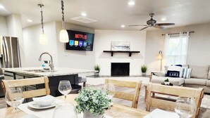 Eine moderne Küche mit integrierter Kochinsel, ein Essbereich mit Holztisch für vier Personen und ein Wohnzimmer mit Sofa und Kamin.