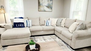 Ein hellbeiges, L-förmiges Sofa mit passendem Hocker, ein kleiner Holztisch mit Pflanze und ein Gemälde an der Wand.