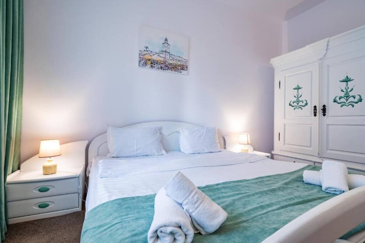 A neatly made bed with white linens and a green blanket, a white wardrobe with decorative handles, and a wall-mounted painting of a town scene.