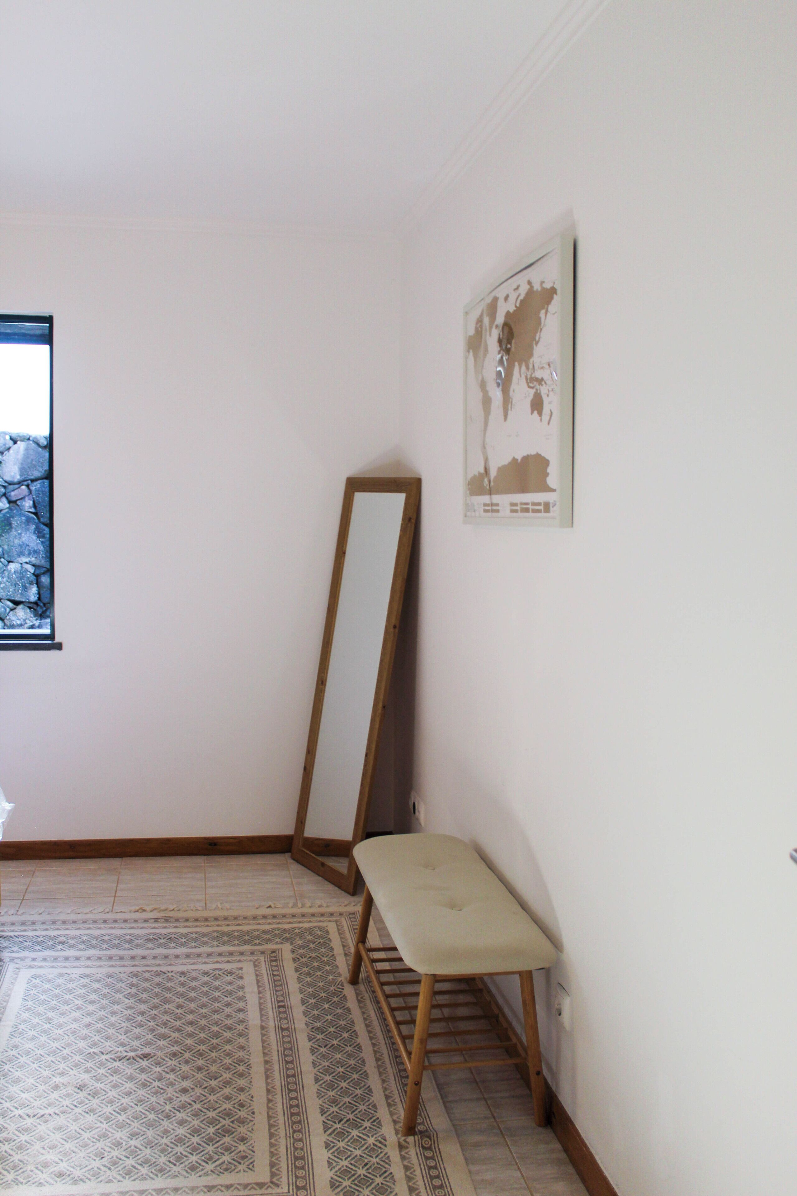 Une pièce avec un tapis à motifs, un banc, un miroir appuyé contre le mur et une carte encadrée.