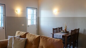 Een gezellige woonkamer met een bank, een eettafel met stoelen en twee ramen met wandlampen.