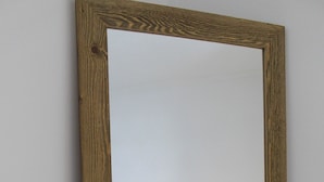 Spiegel met houten lijst die een slaapkamer weerspiegelt, met een bed, nachtkastje en lamp.