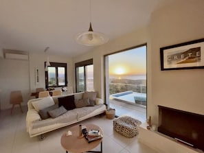 A modern living room with a sofa, coffee table, and fireplace. There is a large window with a view of the sunset.