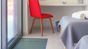 Una stanza con una porta scorrevole in vetro, una sedia rossa, un letto con coperta a quadretti, un tavolino e una finestra vista su del verde.