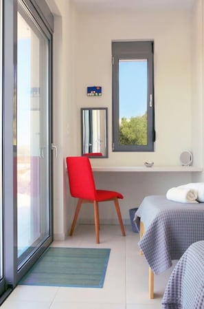 A room with a sliding glass door, a red chair, a bed with a checkered blanket, a small table, and a window with a view of greenery.