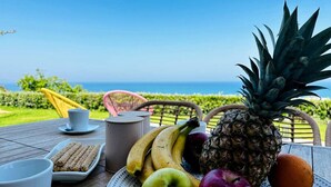 Une table avec des fruits, une tasse et une assiette de nourriture, disposée à l’extérieur, avec vue sur l’océan.