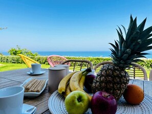 A table with fruits, a cup, and a plate of food, set outdoors with a view of the ocean.