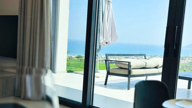 A modern room with a large window overlooking a patio with a bench and umbrella. - Skyline Ocean - 20PL 8BD 6BA, 3min to the beach (GR9M+6V5 Platanias, Greece)