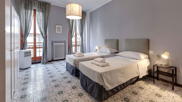 Hotelkamer met twee bedden, een radiator, een elektrische kachel, een nachtkastje met lamp en een raam met gordijnen.