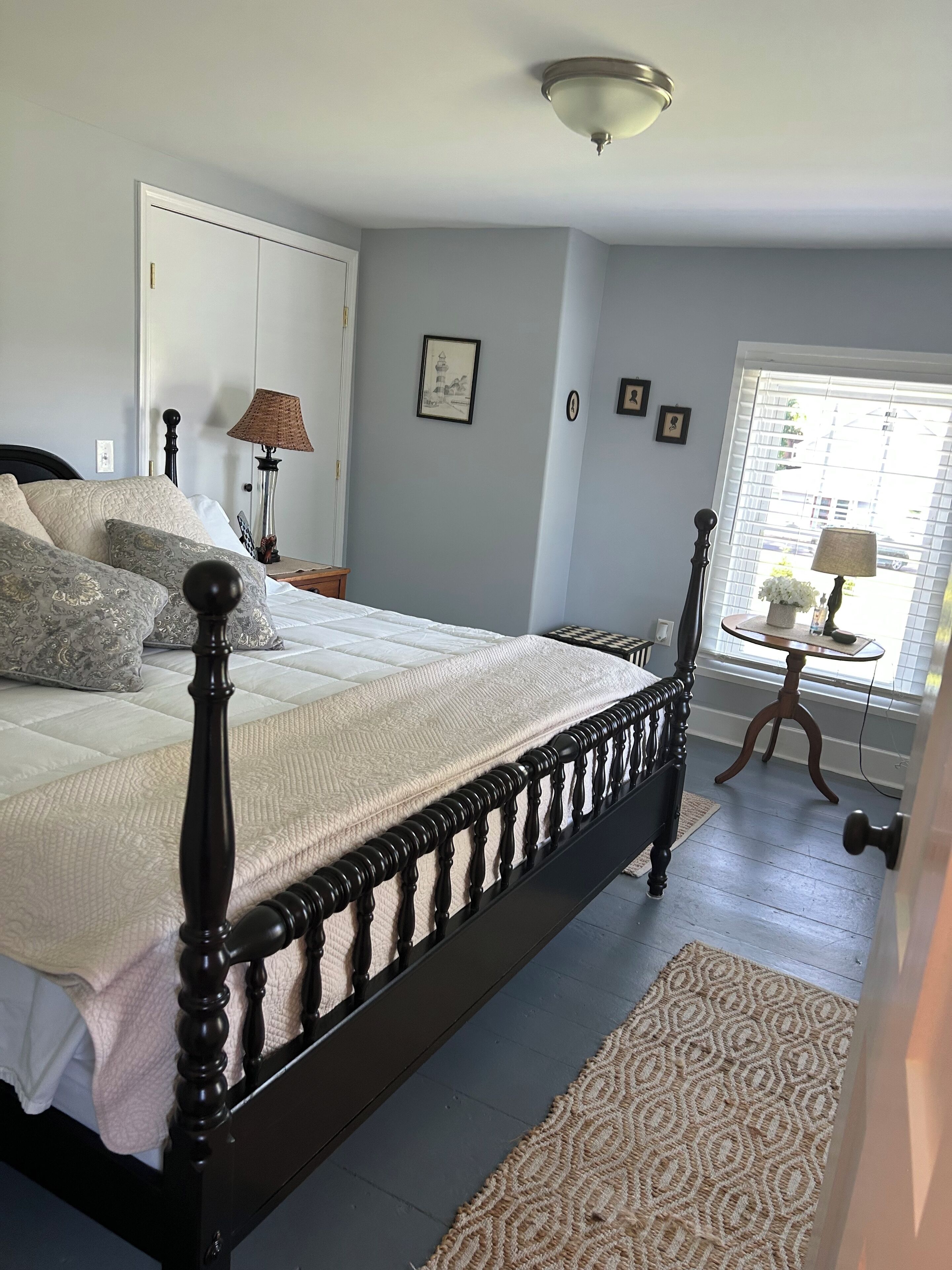 A bedroom with a bed, a nightstand, a lamp, a window with blinds, and a door.