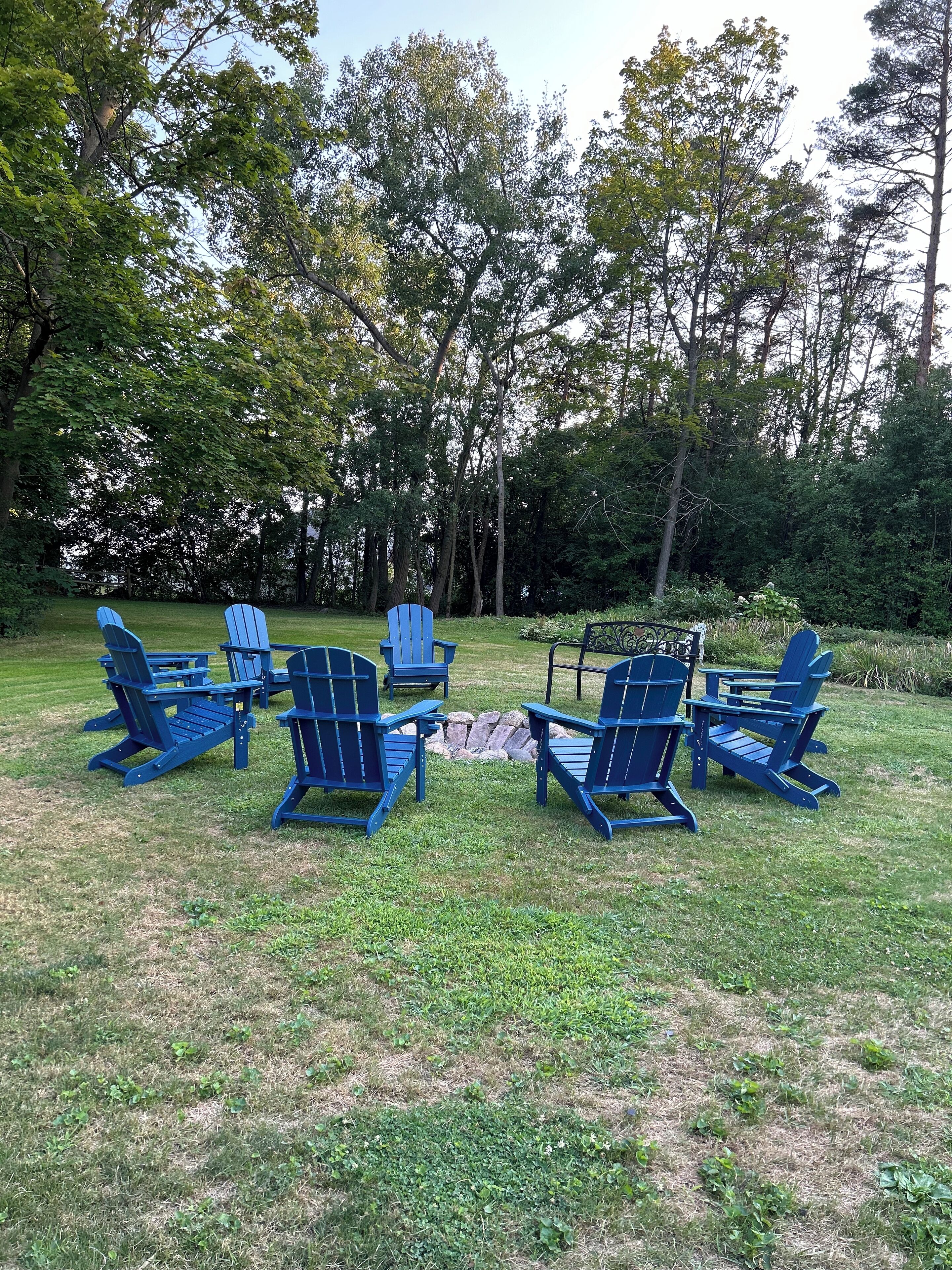 A group of blue Adirondack chairs arranged in a circle around a small fire pit in a grassy outdoor area with trees in the background.