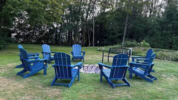 A group of blue Adirondack chairs arranged in a circle around a small fire pit in a grassy outdoor area with trees in the background.