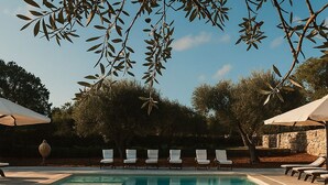 Una piscina rectangular con agua azul cristalina, rodeada de sillas de descanso y sombrillas, con árboles y un cielo despejado de fondo.