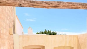 Un patio iluminado por el sol con chimenea de piedra, un jarrón verde y una pequeña zona de estar con sillas y mesa de madera.