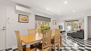 A dining area with a wooden table and chairs, a potted plant, and a view of a brick wall and greenery outside. - Prez Cote La Plaz - Pet Friendly -1 Min Walk to Beach (Callala Bay)