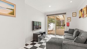 A modern living room with a black and white checkered floor, a grey sofa, a flat-screen TV, and a sliding glass door leading to an outdoor area. - Prez Cote La Plaz - Pet Friendly -1 Min Walk to Beach (Callala Bay)