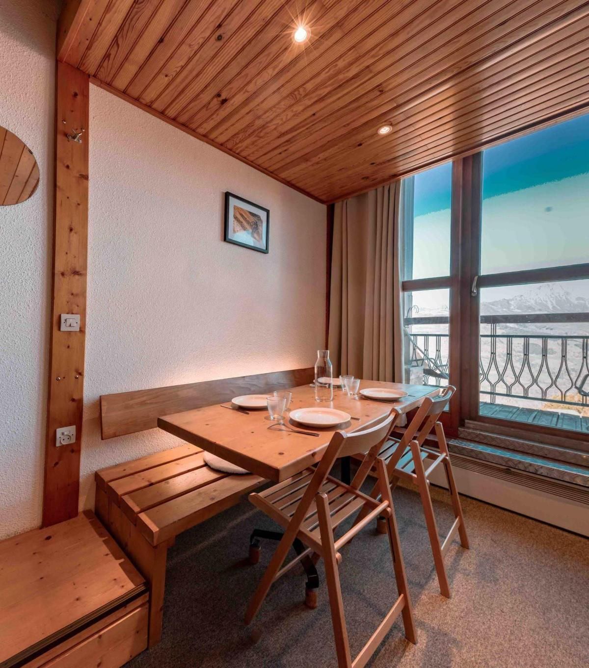 A wooden interior with a table set for two, a bench, and a window with a view of mountains.