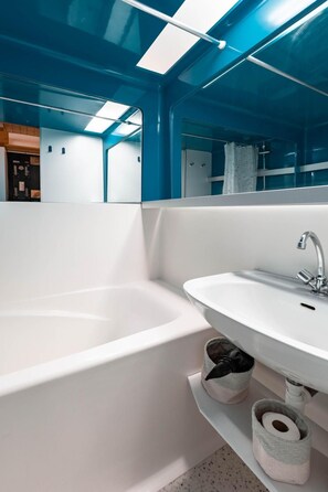 Ein modernes Badezimmer mit einer weißen Badewanne, einem Waschbecken und einem Spiegel. Unter dem Waschbecken befinden sich zwei Körbe, einer mit einem schwarzen Gegenstand und der andere mit einer Toilettenpapierrolle.