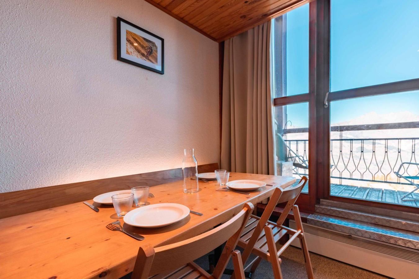 A dining table with chairs, plates, glasses, and cutlery. There is a window with a view of mountains and a balcony.
