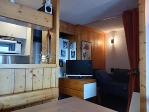 Quarto compacto com paredes de madeira, uma kitchenette com micro-ondas, uma TV sobre um móvel de madeira e uma cama com cobertura escura.