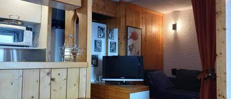 A compact room with wooden walls, a kitchenette with a microwave, a TV on a wooden cabinet, and a bed with a dark cover.