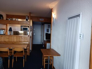 Een compacte keuken met houten kastjes, een magnetron en een spoelbak. Er is een eethoek met een tafel en stoelen.