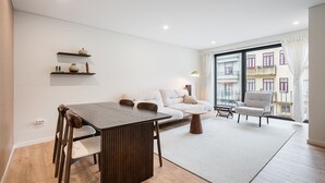 A modern living room with a wooden dining table, a sofa, and a chair. There is a large window with a view of buildings.