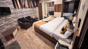 Une chambre d’hôtel moderne avec un mur en pierre, un lit recouvert de draps blancs, un bureau en bois, une chaise, un téléviseur fixé au mur et un miroir.
