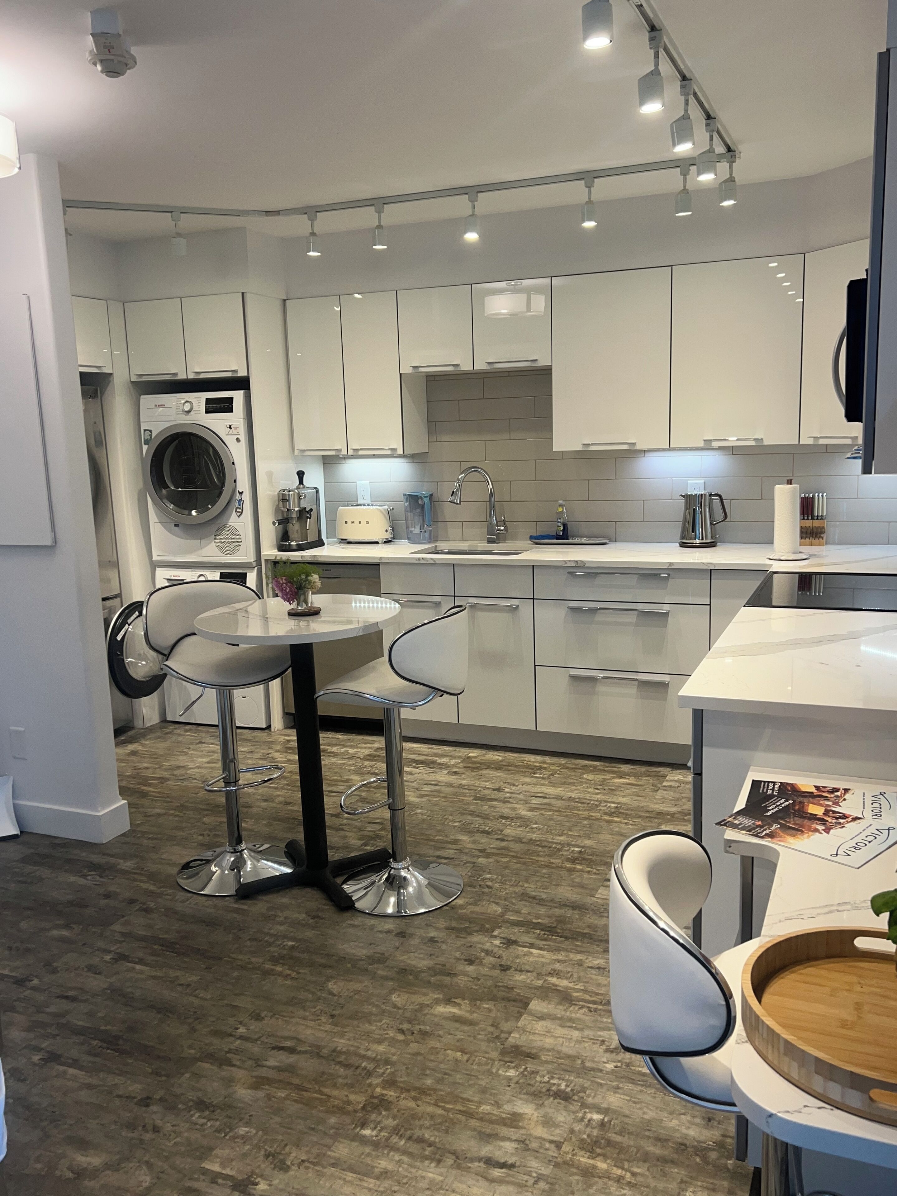 A modern kitchen with white cabinets, a built-in washing machine, a dining area with a round table and two bar stools, and a wooden floor.