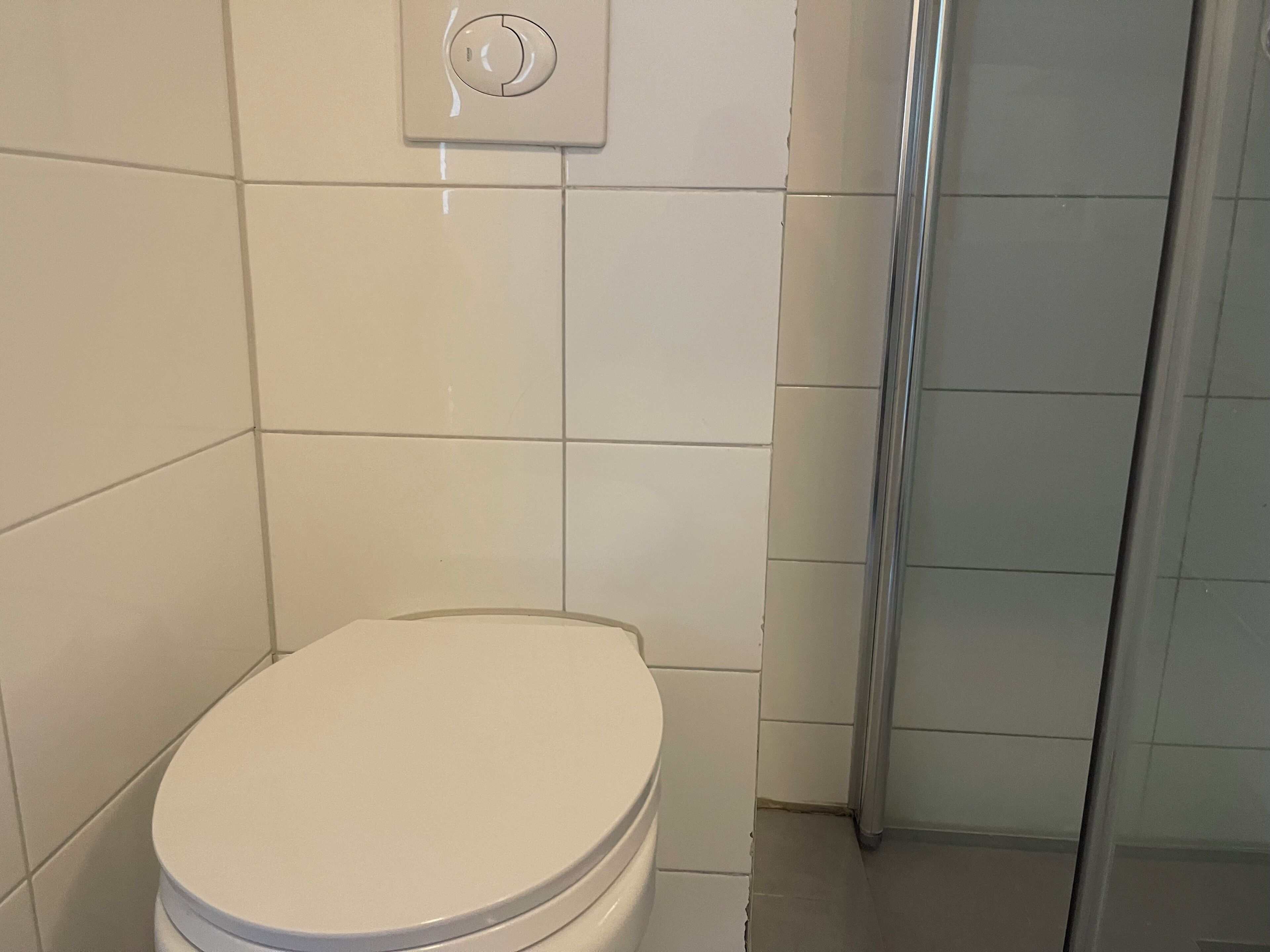 Des toilettes avec un abattant blanc, une poignée de chasse d’eau et un mur recouvert de carreaux.
