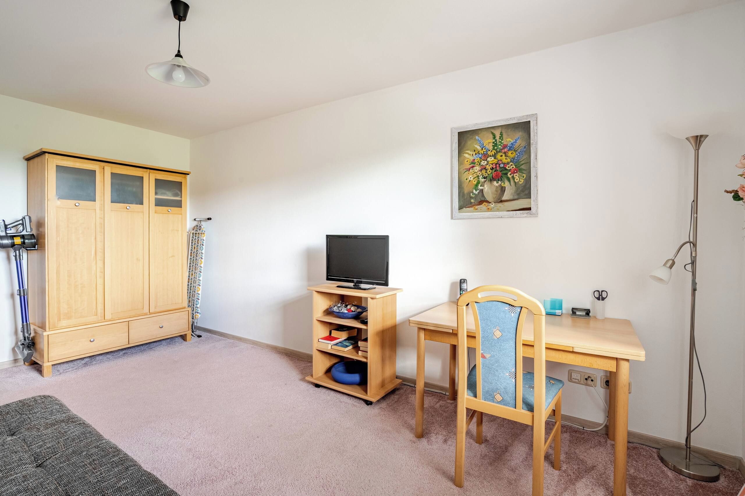 Quarto com armário de madeira, televisão em suporte, escrivaninha com cadeira e abajur.
