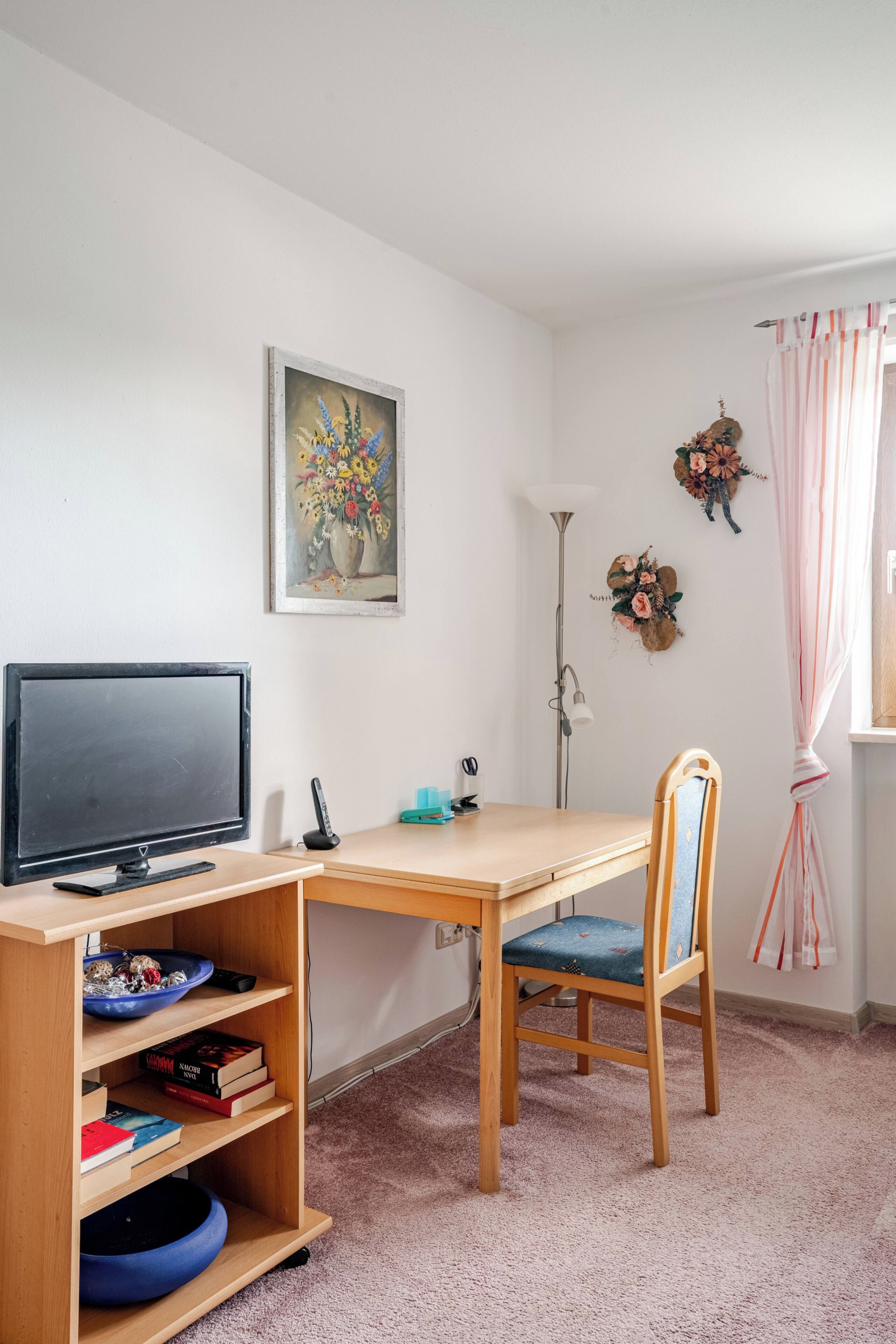 Huoneessa on puinen työpöytä, tuoli, televisio, kirjahylly ja seinällä taulu.