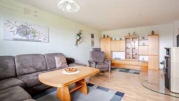 A modern living room with a grey sofa, a wooden coffee table, and a grey armchair. There is a kitchen area with cabinets and a refrigerator.