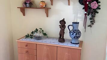 Puisesta valmistettu kaappi, jossa on laatikoita, ja sen päällä on pitsinen pöytäliina. Kaappia koristavat keramiinen kannu, pieni koristehahmo ja maljakko, jossa on kukkia.