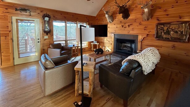 A cozy cabin interior with a fireplace, wooden walls, a ceiling fan, and deer antlers decor.