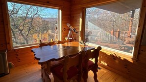 A wooden cabin with a dining table and chairs, large windows, and a telescope.