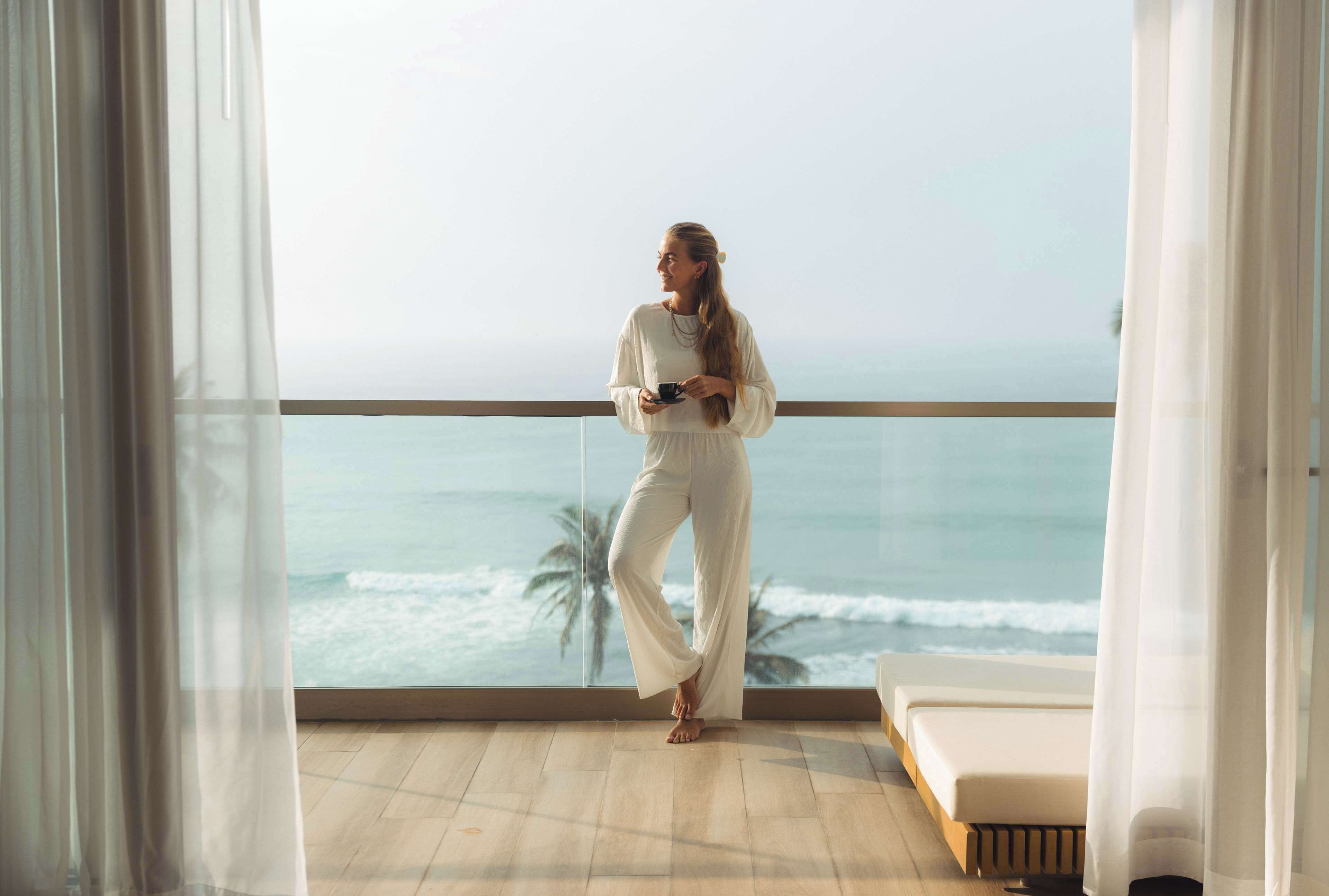A room with a large window overlooking the ocean, a bed, and a balcony with a railing.