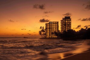 A beachfront hotel with a modern design, featuring multiple floors and balconies, located near the ocean with waves crashing onto the shore.