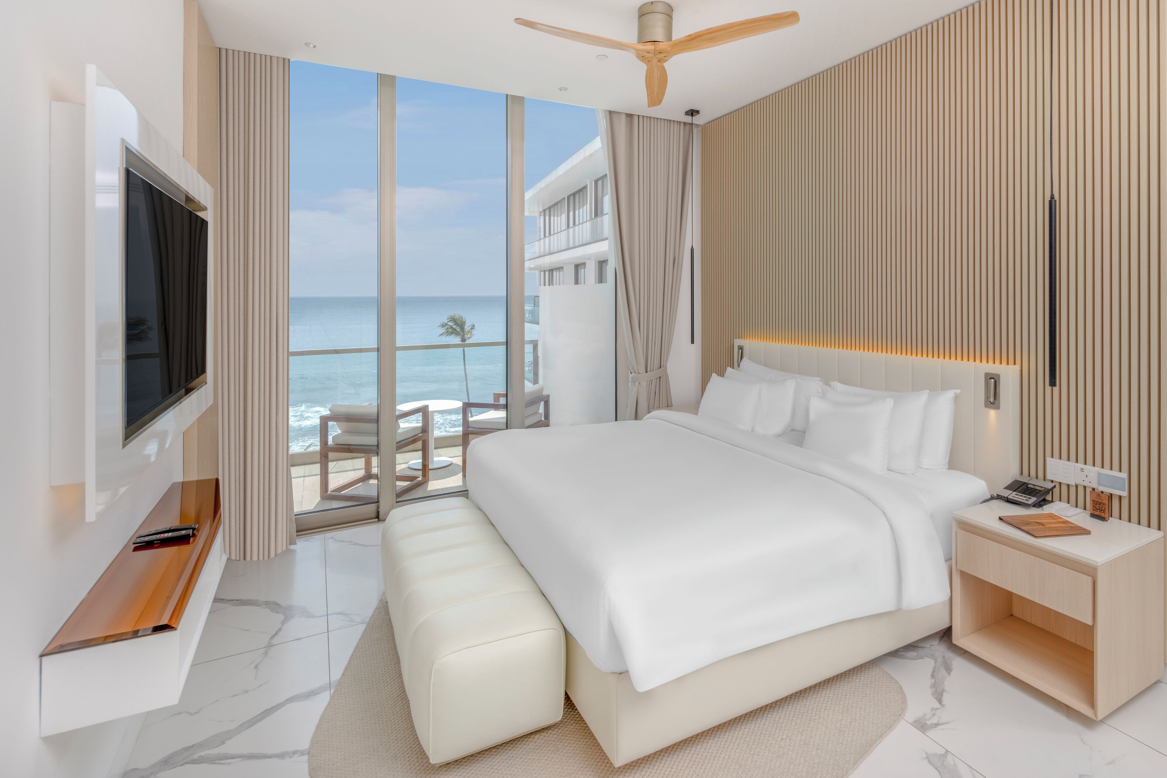 Una camera d'albergo moderna con un letto grande, una TV, un comodino in legno e un balcone con vista sull'oceano.