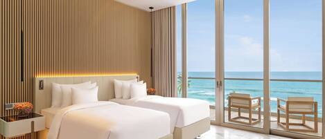 A modern hotel room with two beds, a ceiling fan, a large window with a view of the ocean, and a balcony with seating.
