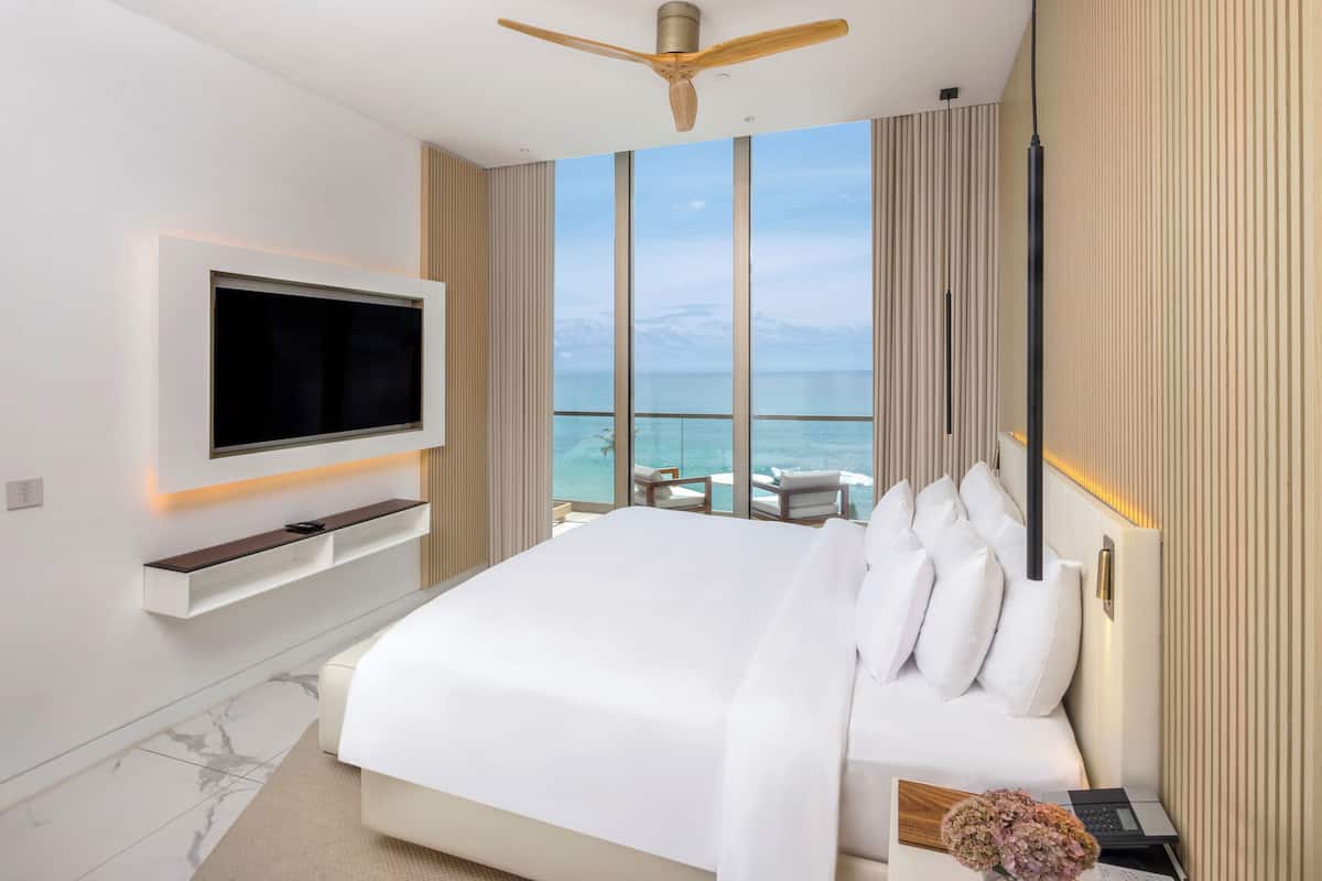 Une chambre d’hôtel moderne dotée d’un grand lit, d’un téléviseur fixé au mur et d’un balcon offrant une vue sur l’océan.