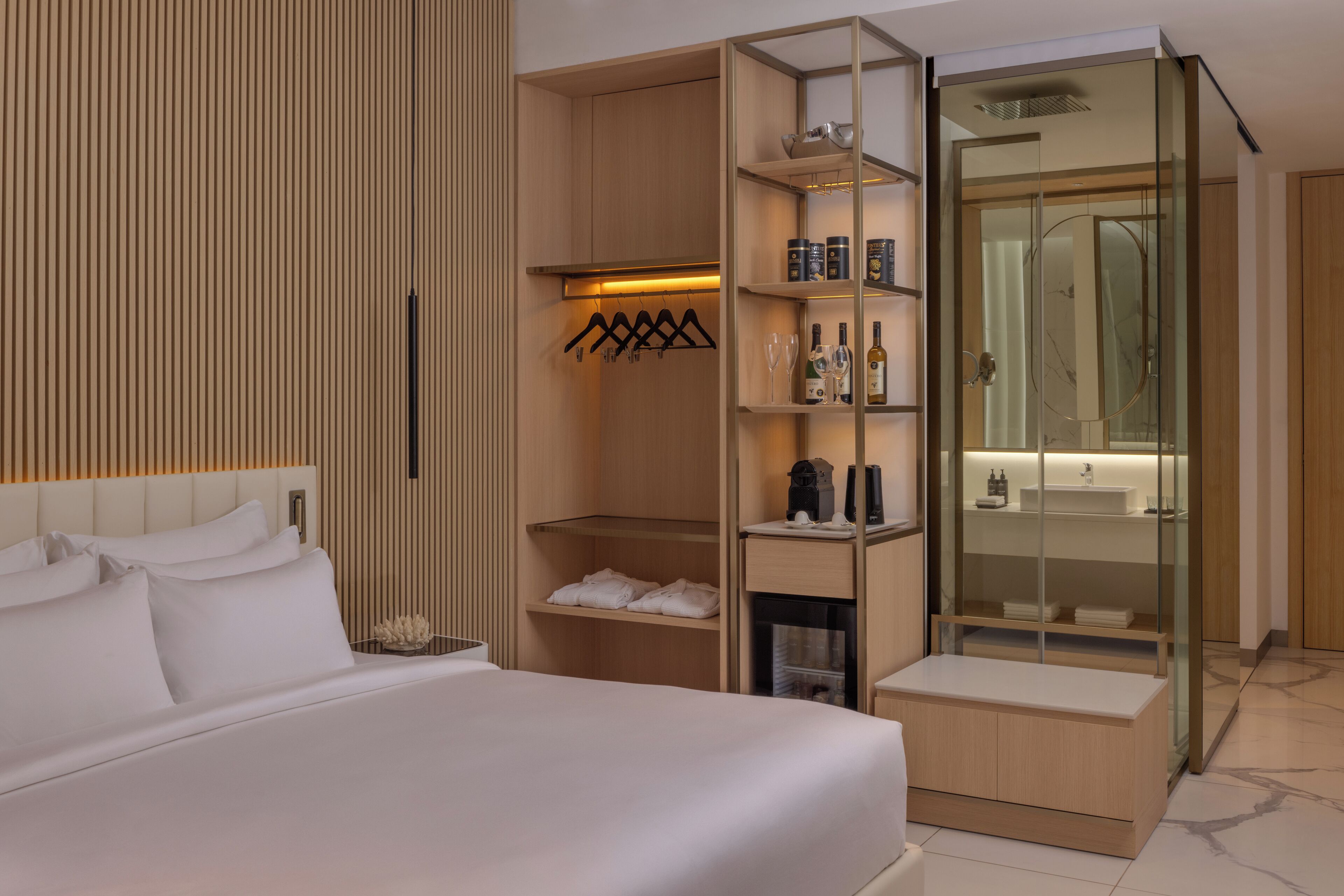 A modern hotel room with a large bed, a walk-in closet, a glass shower enclosure, and a vanity with a mirror.