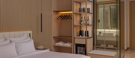 A modern hotel room with a large bed, a walk-in closet, a glass shower enclosure, and a vanity with a mirror.