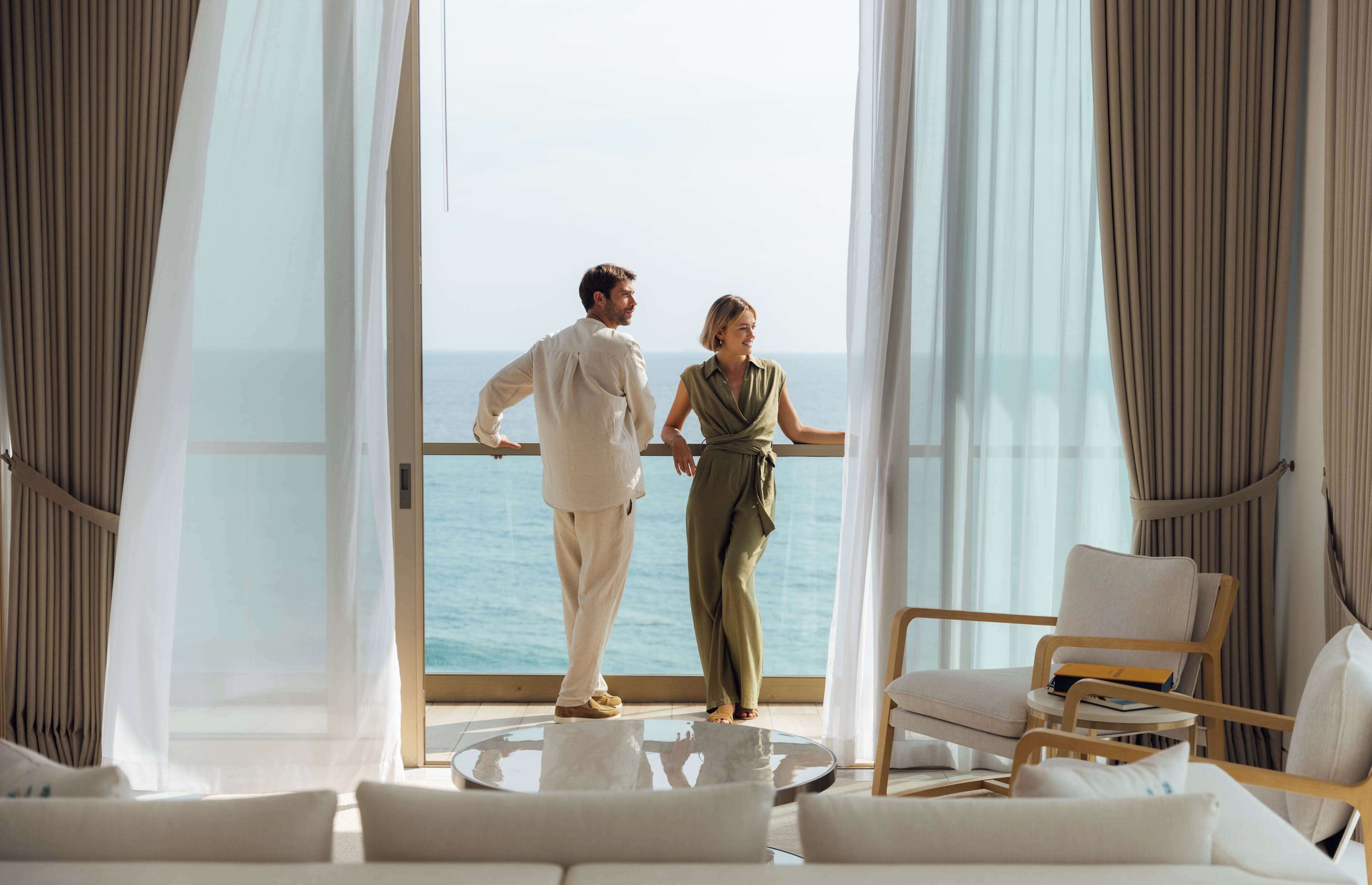Una coppia in piedi vicino a una grande finestra con vista sul mare, in un ambiente moderno che include un tavolo rotondo in vetro e posti a sedere confortevoli.