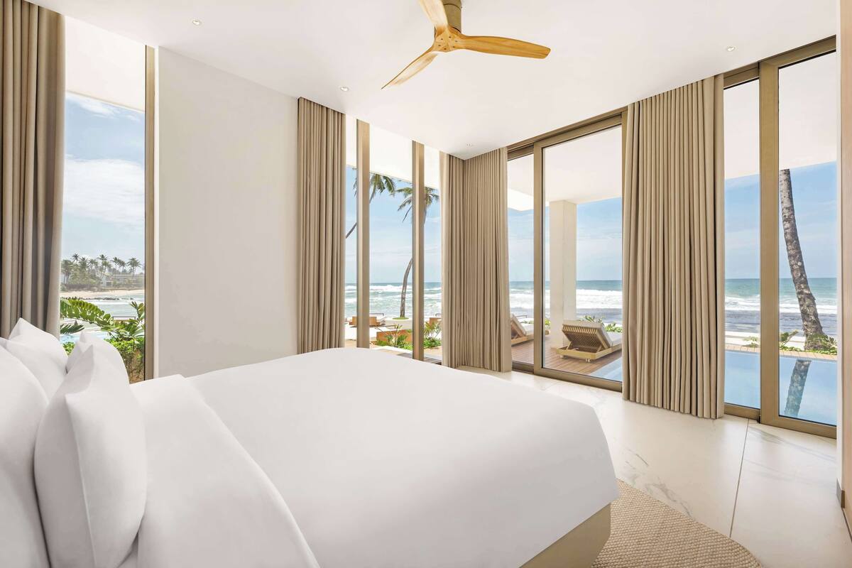 Une chambre d’hôtel moderne avec un grand lit, un ventilateur de plafond et une vue sur l’océan et les palmiers.