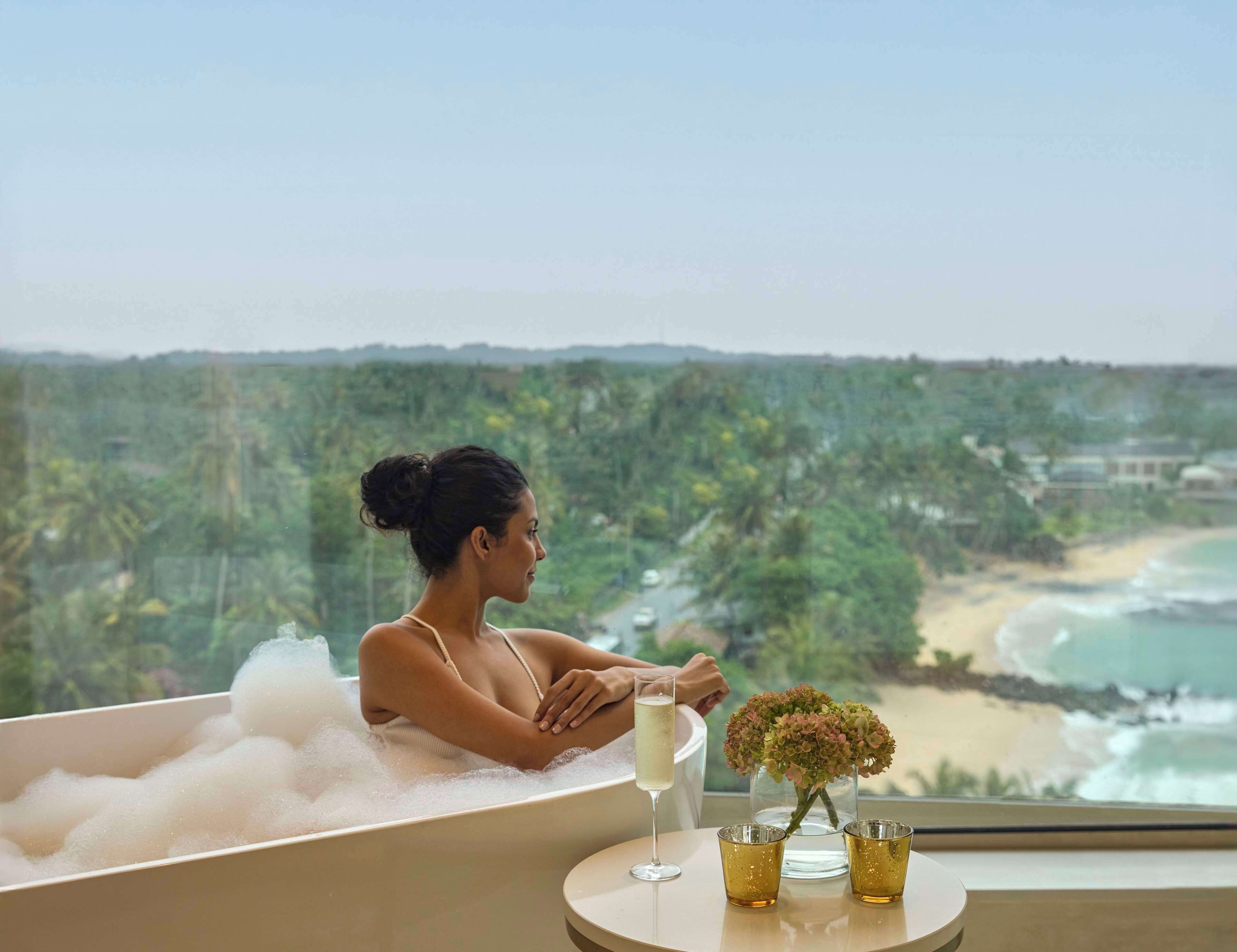 A woman relaxing in a bathtub with a view of a beach and forest. There are two glasses and a vase with flowers on a table beside her.