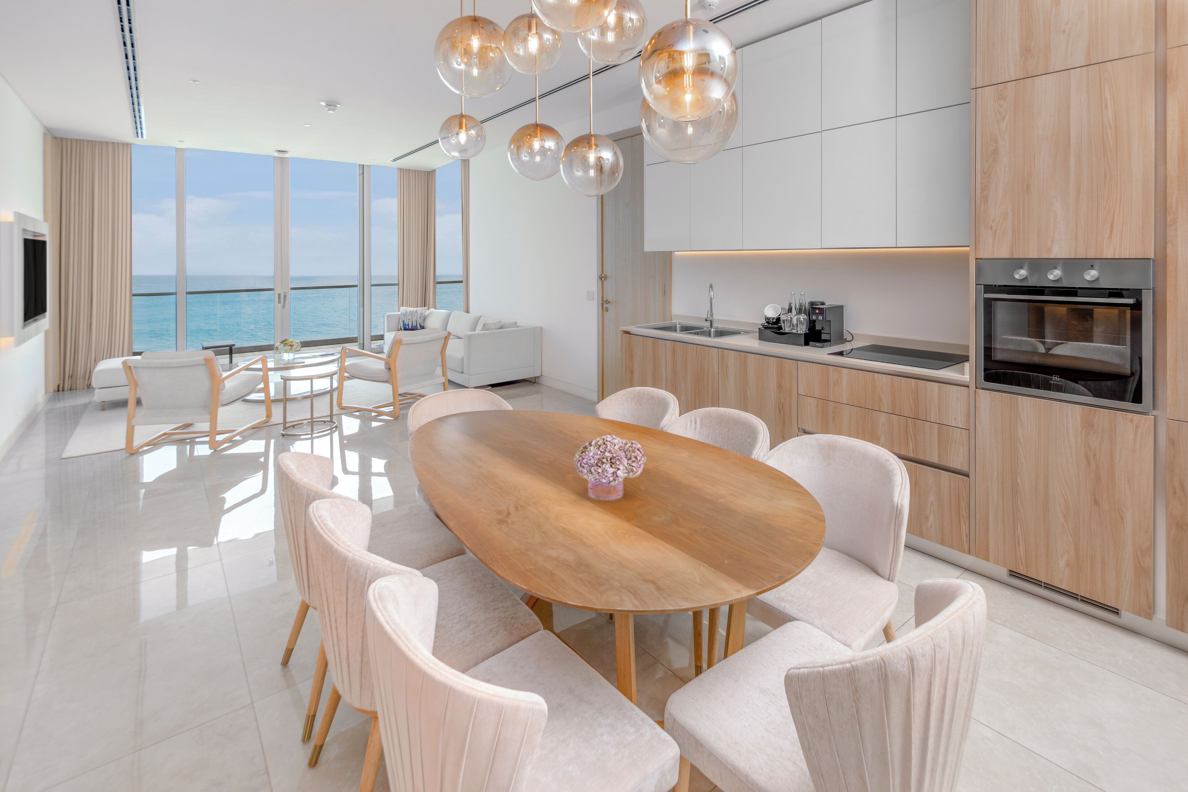 A modern kitchen with a large island, a dining area with a wooden table and white chairs, and a view of the sea.