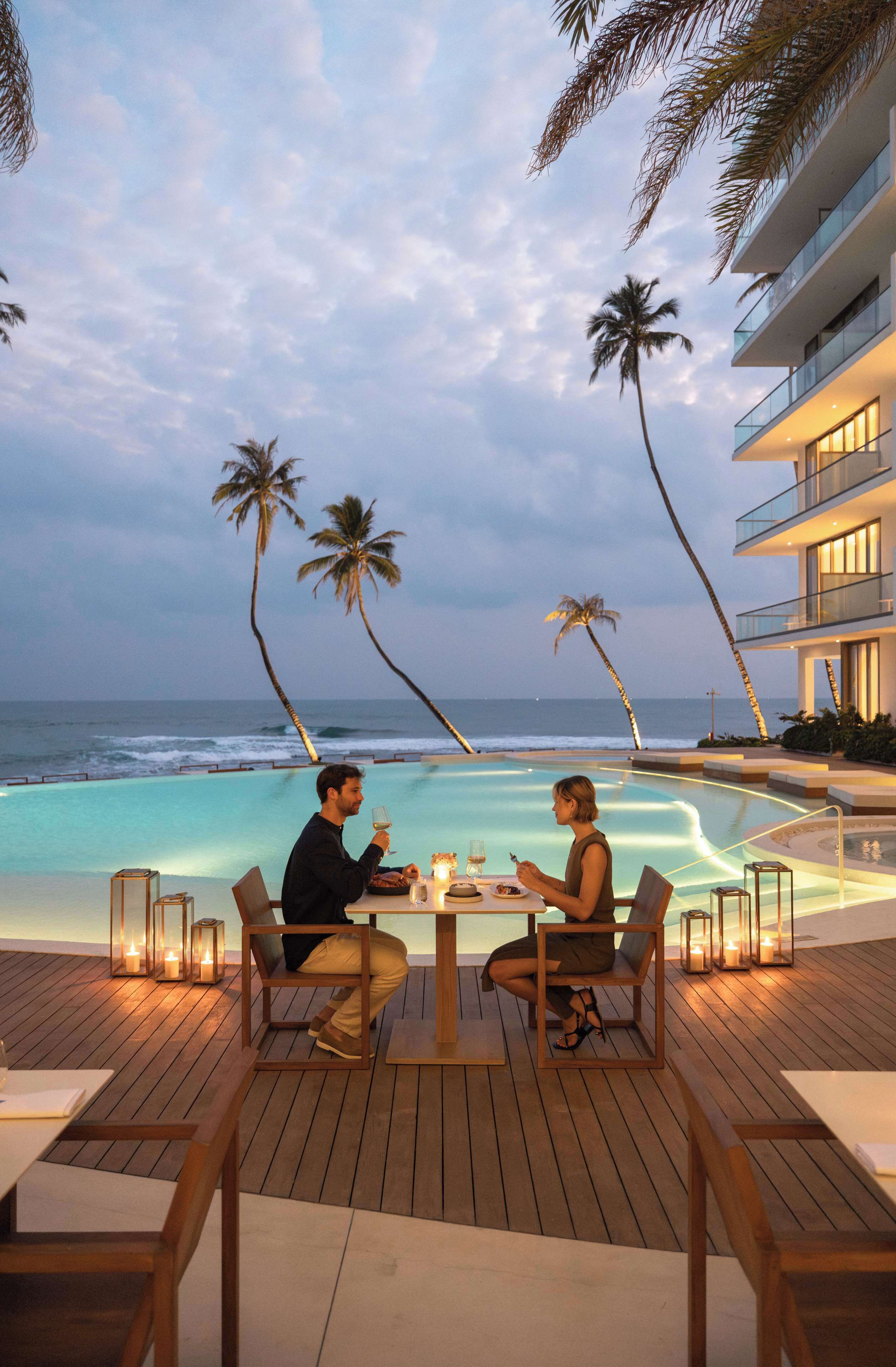 A couple dining at a wooden deck with a view of the ocean and palm trees.